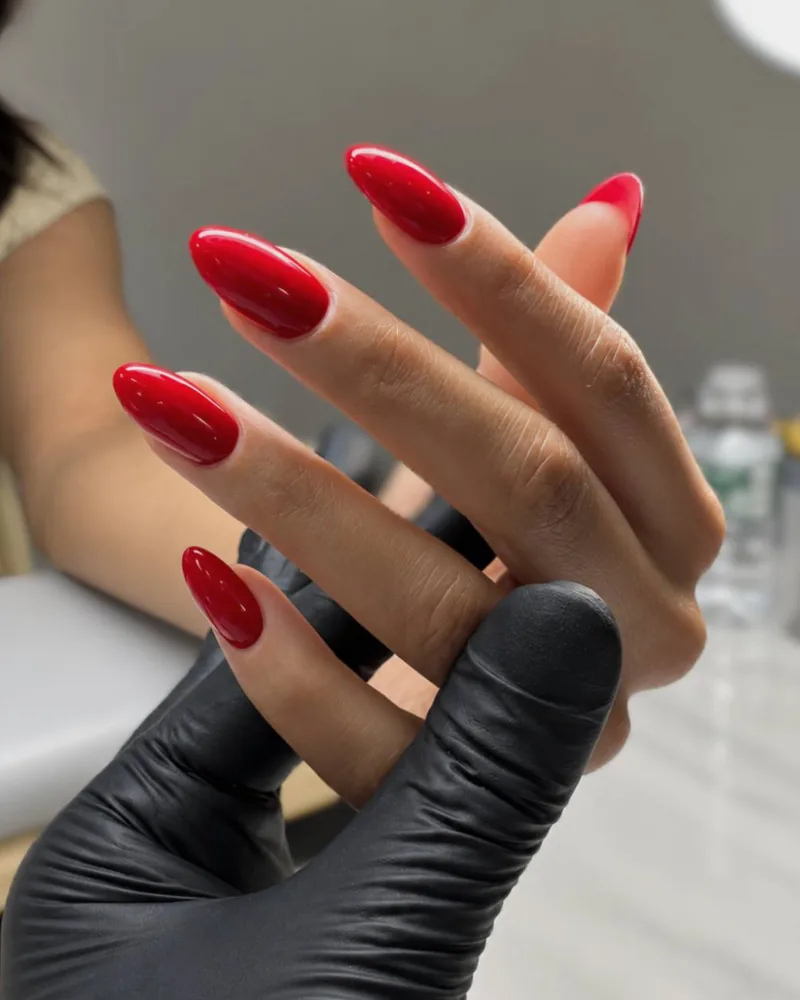 The nail tech's gloved hand holding a client's hand with red almond nails