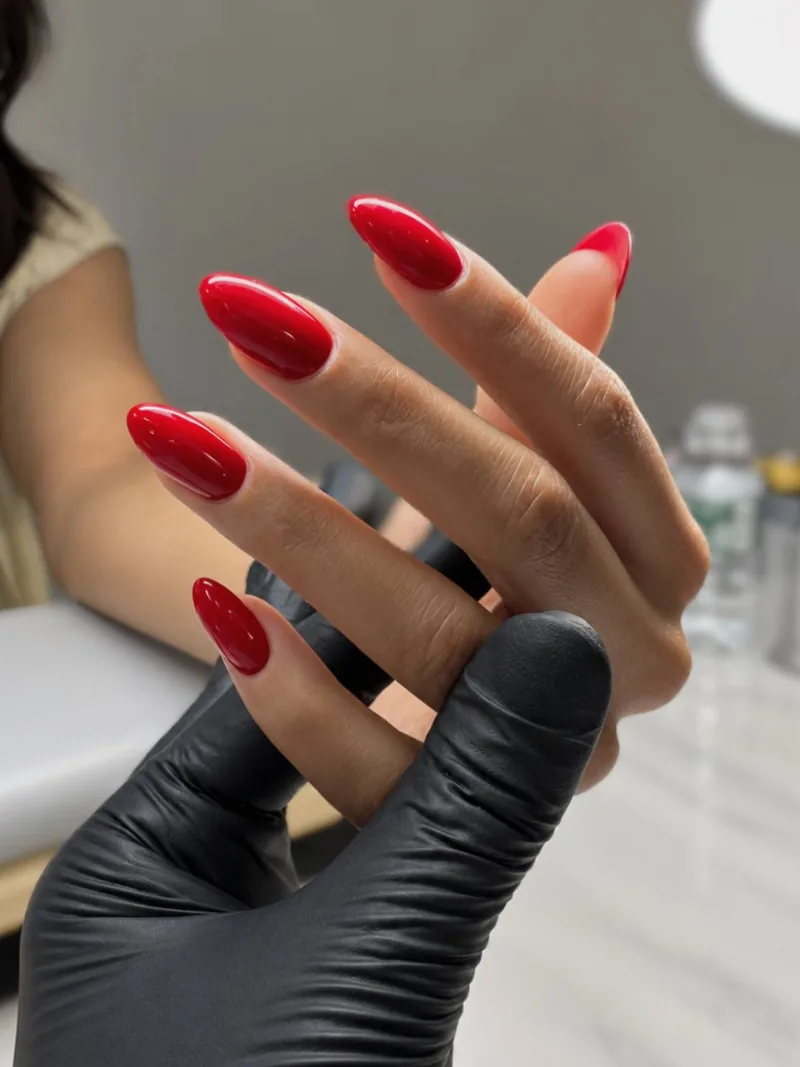 Almond nails in glossy classic red gel, held in the nail tech's gloved hand